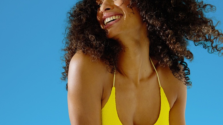 Smiling woman in a yellow swimsuit with curly hair, looking to the side against a bright blue background.