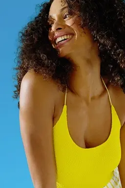 Smiling woman in a yellow swimsuit with curly hair, looking to the side against a bright blue background.