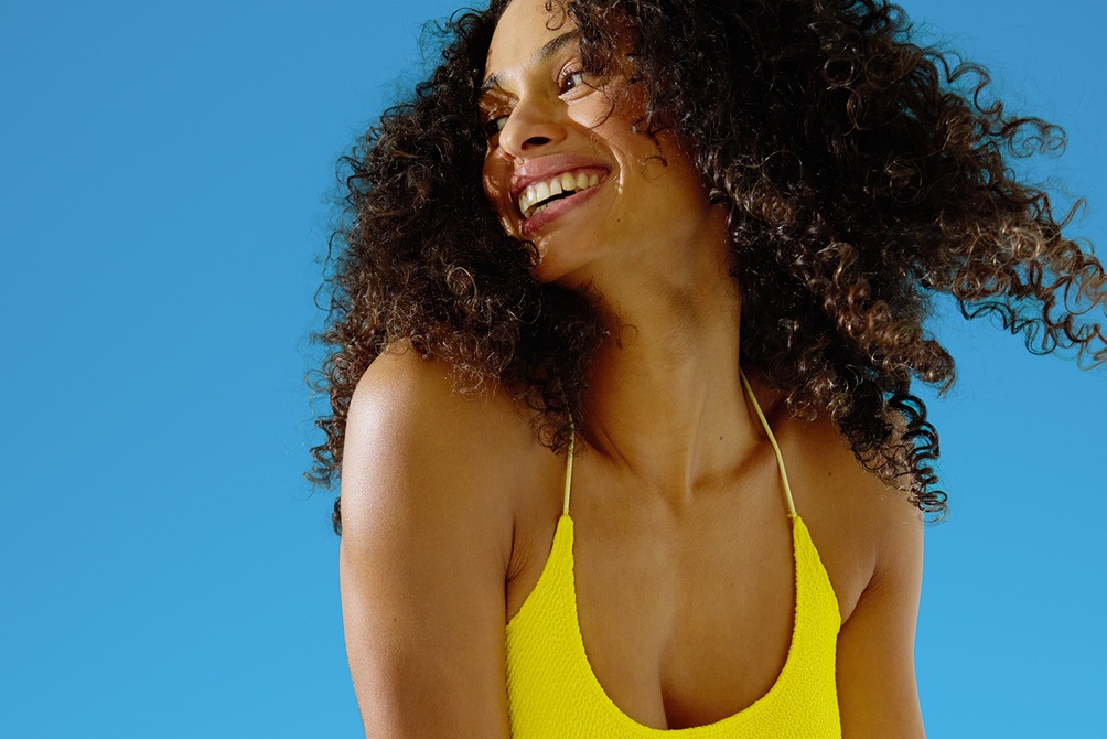Smiling woman in a yellow swimsuit with curly hair, looking to the side against a bright blue background.