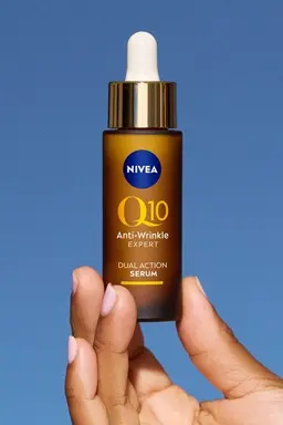 Hand holding a brown dropper bottle of NIVEA Q10 Anti-Wrinkle Expert Dual Action Serum against a blue background