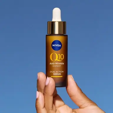 Hand holding a brown dropper bottle of NIVEA Q10 Anti-Wrinkle Expert Dual Action Serum against a blue background