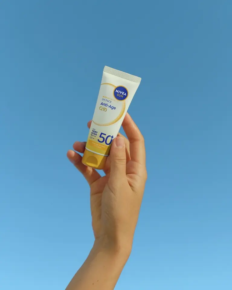 Hand holding a tube of NIVEA Sun UV Face Anti-Age Q10 sunscreen with SPF 50+ against a blue background