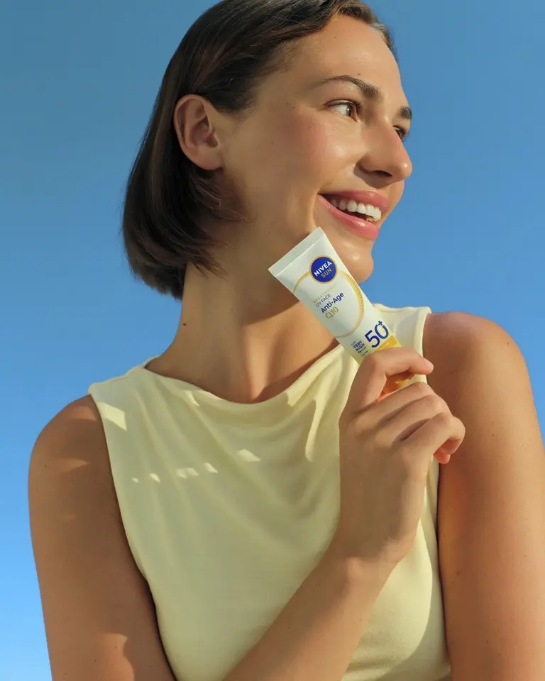 Smiling woman holding a tube of NIVEA Sun UV Face Anti-Age Q10 SPF 50 beside her cheek against blue