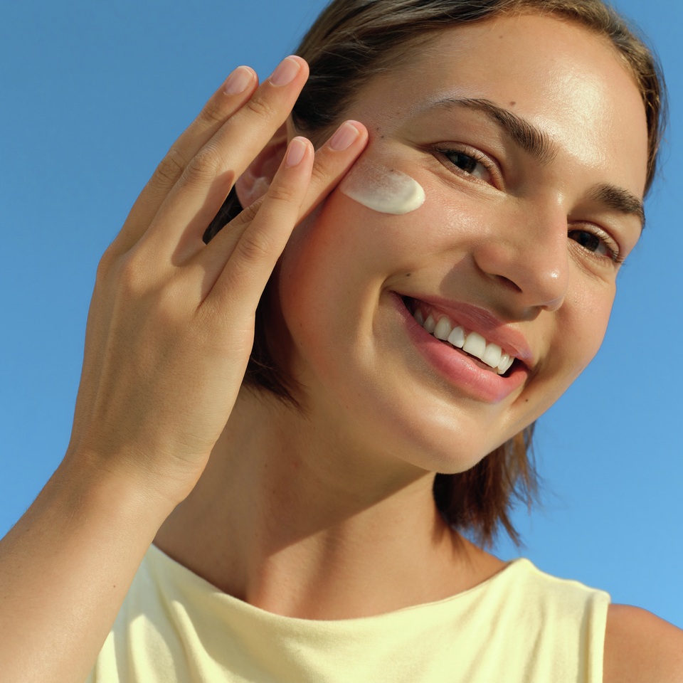 Smiling woman applying white face cream to her cheek with her fingertips against a clear blue sky