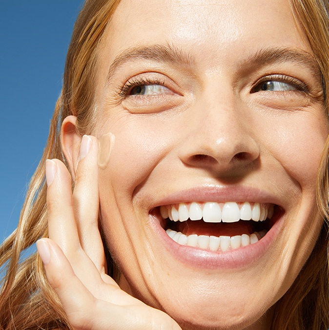 A woman smiles while swiping on daily fluid to her cheek.