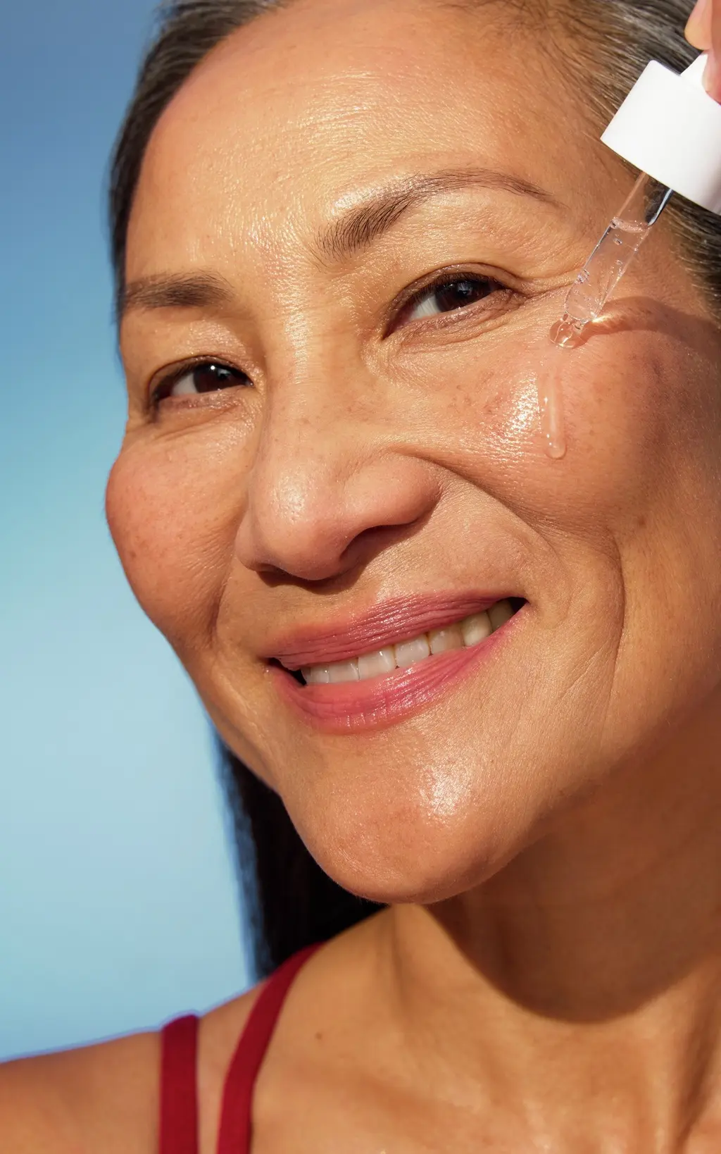 Smiling mature woman applying clear facial serum to her cheek with a glass dropper against a blue background