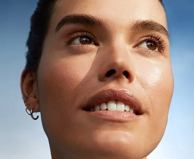 Close-up of a woman looking upward in sunlight, with dewy skin against a clear blue sky
