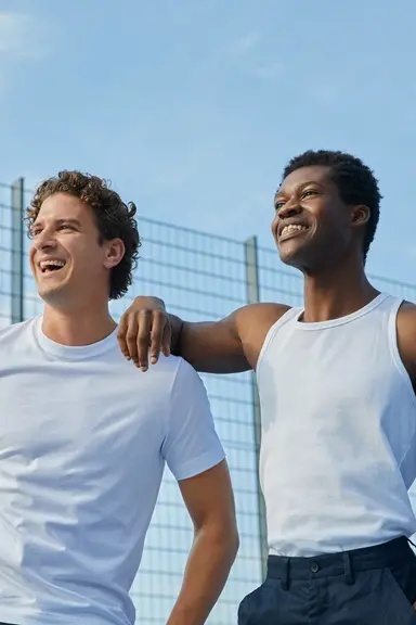 Two smiling men in white tops standing arm in arm beside an outdoor sports court fence