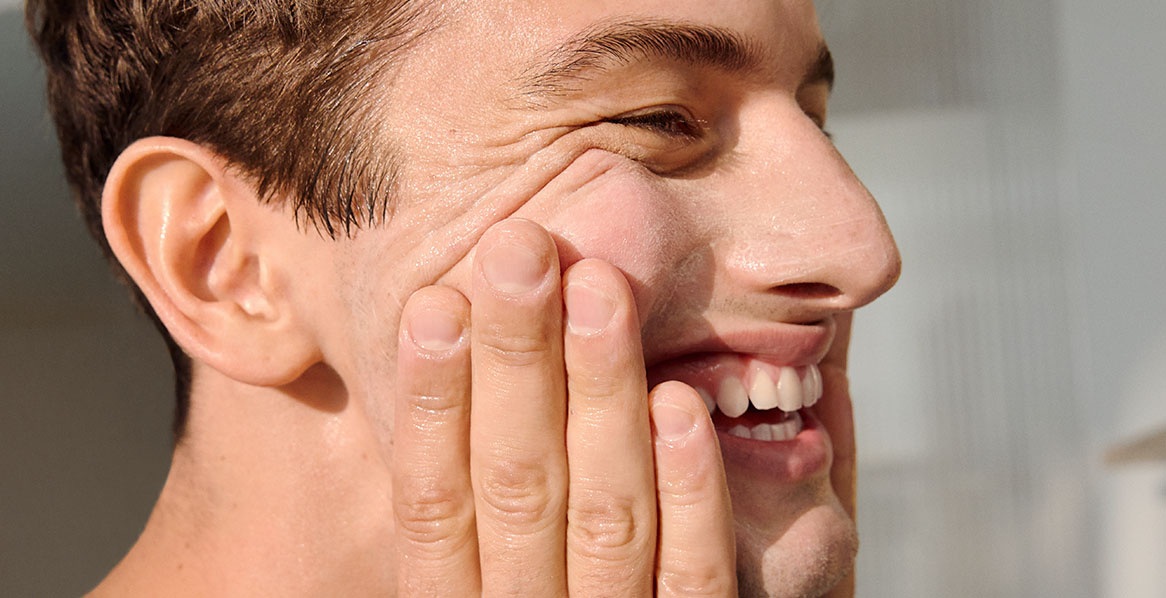 A side profile of a young grinning man with radiant skin, pressing his hands to his cheeks.