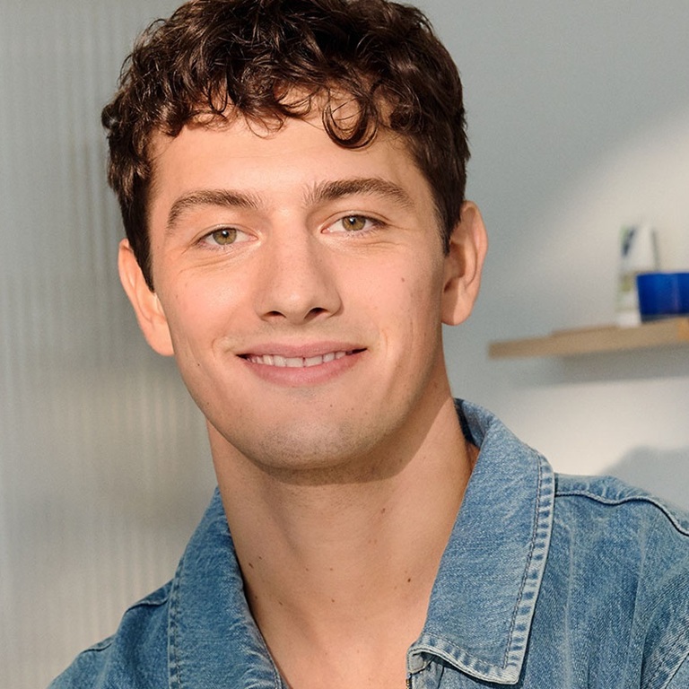 A young man with radiant facial skin casually leans and smiles in a bathroom setting, lifting an eyebrow.