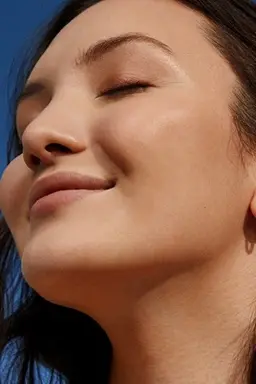 A young woman smiles with eyes closed, showing the glowing skin of her face and neck.