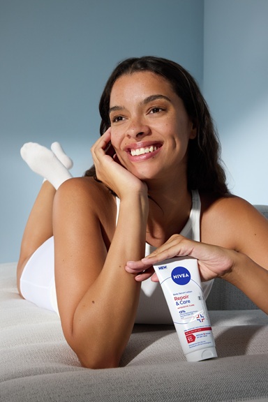 Smiling woman reclining on a sofa while holding a tube of NIVEA Repair & Care body serum lotion
