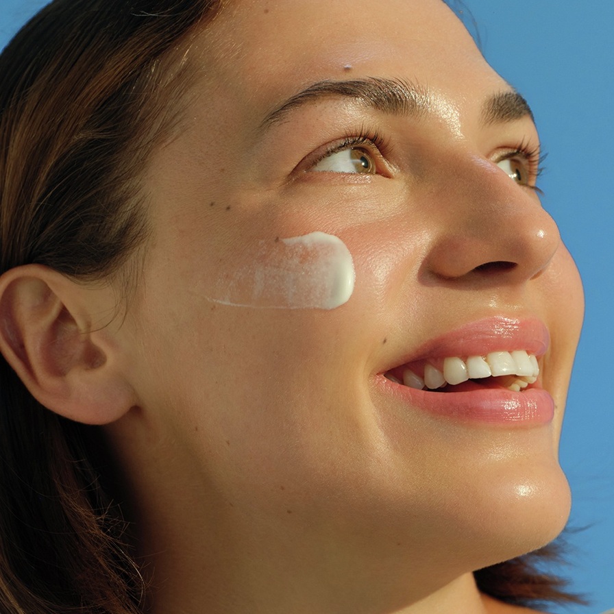 A young woman smiles and gazes upwards with a swipe of sunscreen on her glowing cheek.